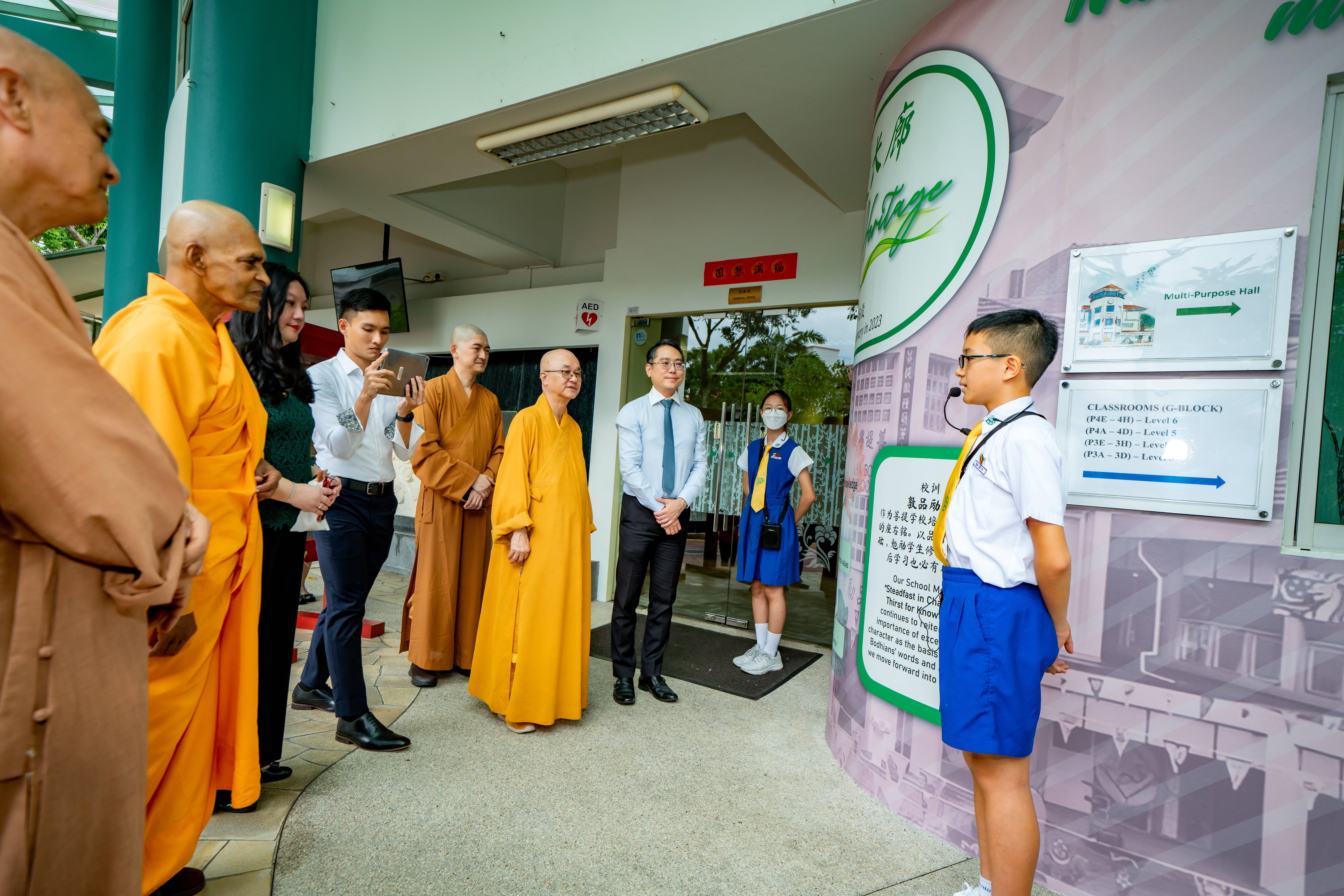 A young student with a microphone speaks to a group of Buddhist monks and adults at a school entrance.